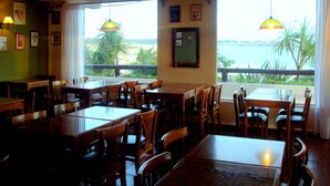 A restaurant with wooden tables and chairs, large windows with a view of the sea, and hanging lamps. - Hotel Morgan Bay (Piriapolis)