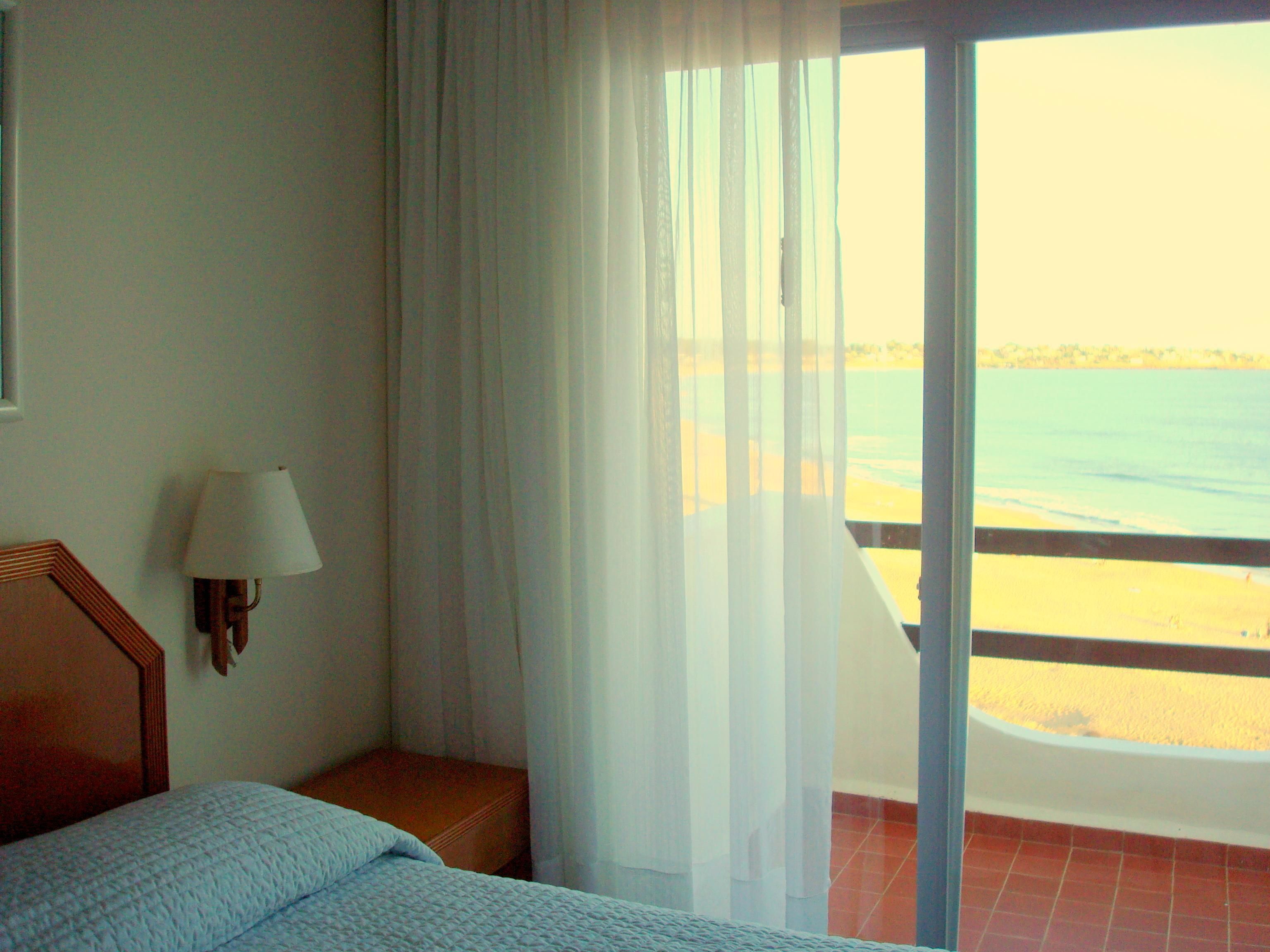 A hotel room with a bed, a nightstand, a lamp, and a view of the beach through a large window.