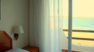 A hotel room with a bed, a nightstand, a lamp, and a view of the beach through a large window.