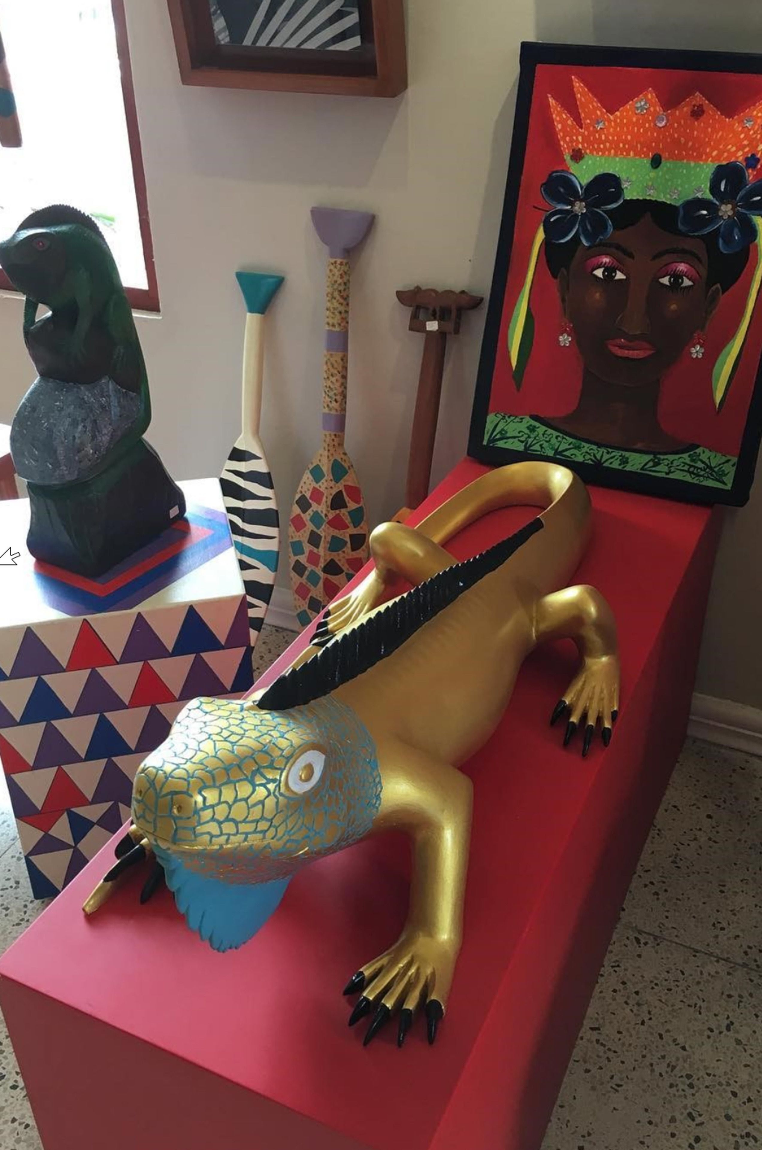 Una escultura colorida de un lagarto sobre un pedestal rojo, rodeada de diversos objetos decorativos, incluyendo un cuadro enmarcado y una escultura verde.