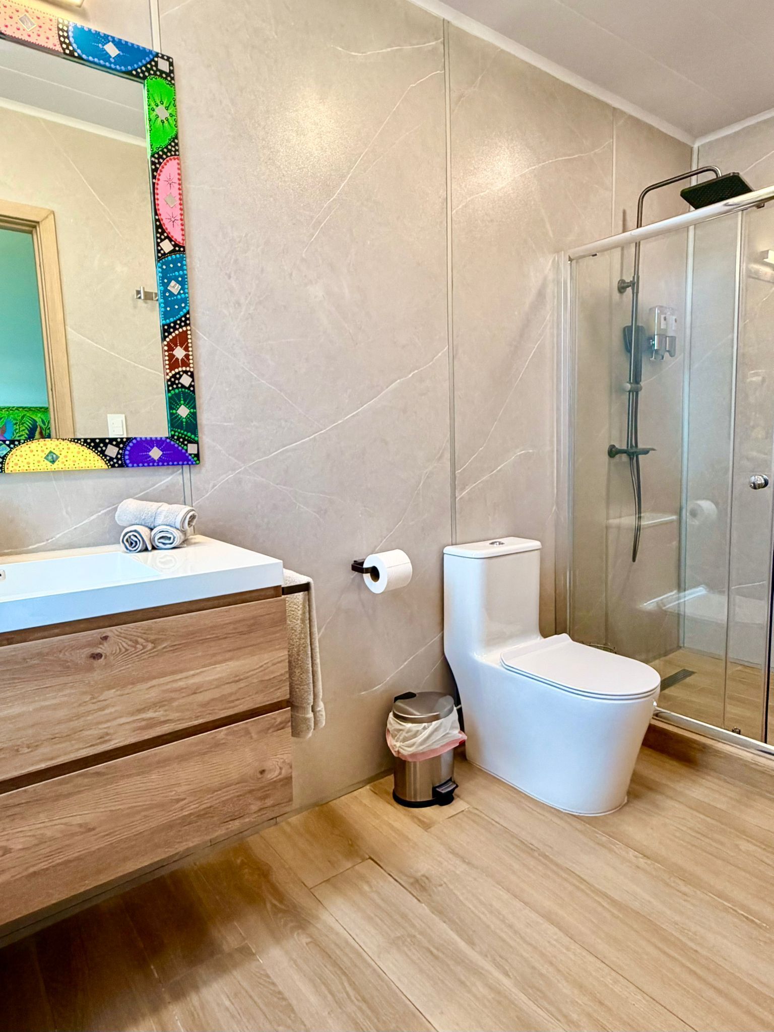A bathroom with a wooden vanity, a toilet, a shower enclosure, and a mirror with a colorful frame.