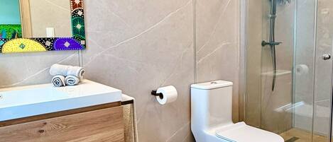A bathroom with a wooden vanity, a toilet, a shower enclosure, and a mirror with a colorful frame.