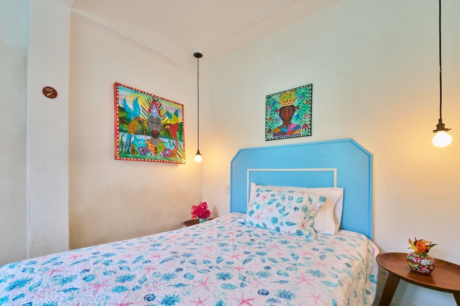 A bedroom with a blue headboard, floral bedspread, two wall-mounted artworks, a small table with a vase, and a hanging light fixture.