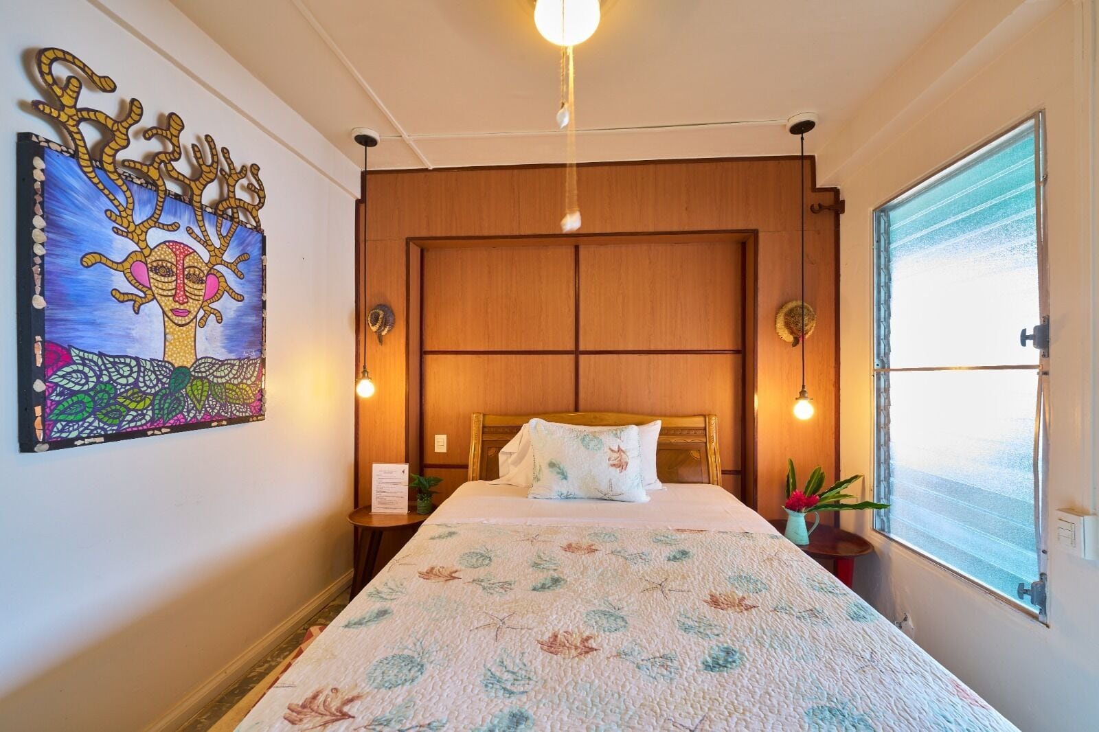 Ein Hotelzimmer mit einem Bett, einem Wandbild, einem Fenster und einem Nachttisch mit einer Lampe.