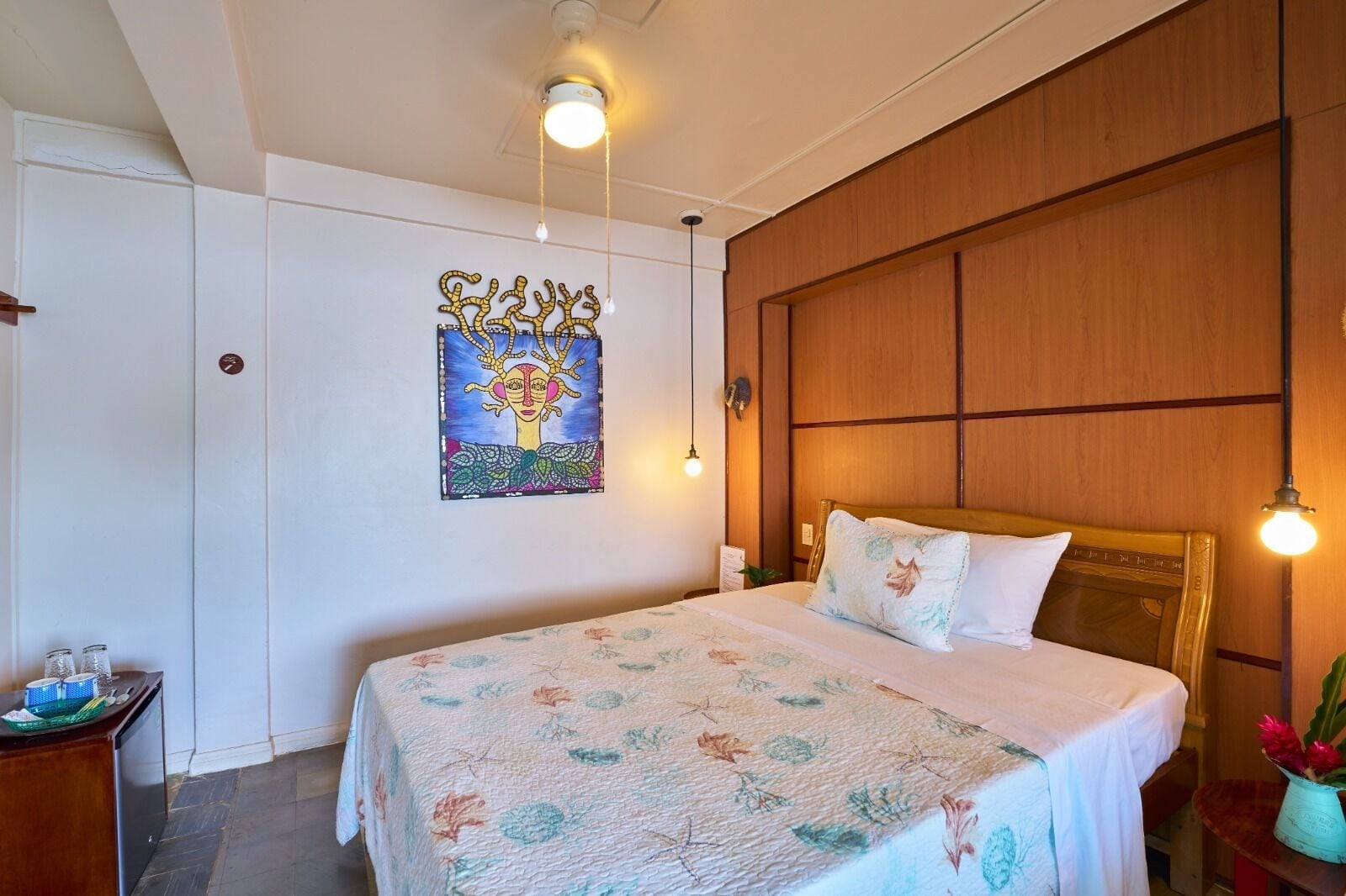 A bedroom with a wooden headboard, a floral bedspread, a wall art piece, and a bedside table with a lamp.