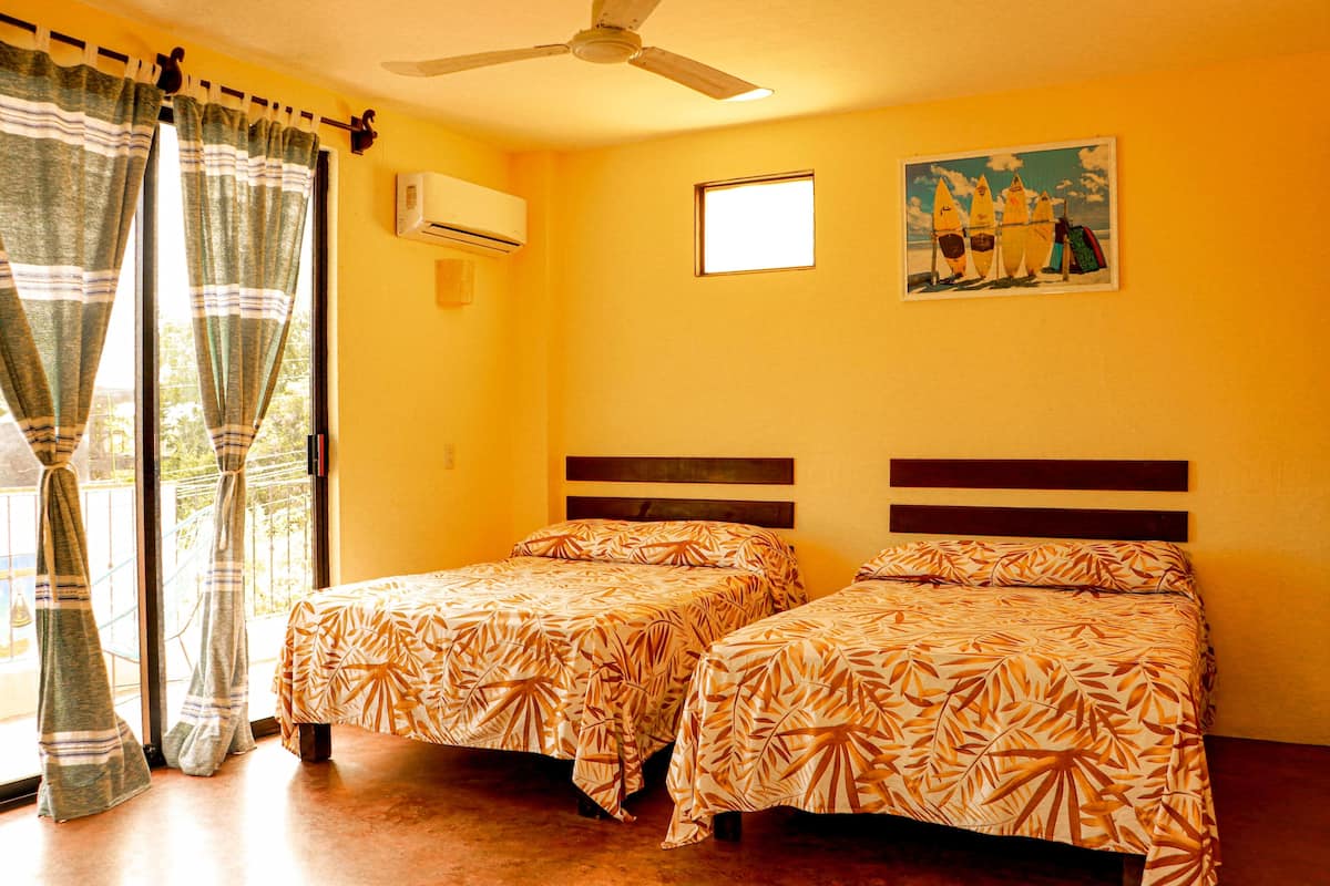 Una habitación con dos camas, un ventilador de techo, una ventana con cortinas, un equipo de aire acondicionado y un cuadro en la pared.
