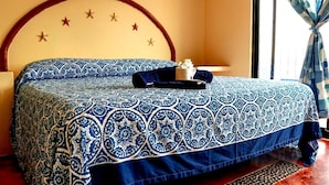A bed with a blue and white patterned bedspread, a wooden headboard with star decorations, and a window with blue curtains. - Rivera del Mar (Puerto Escondido)