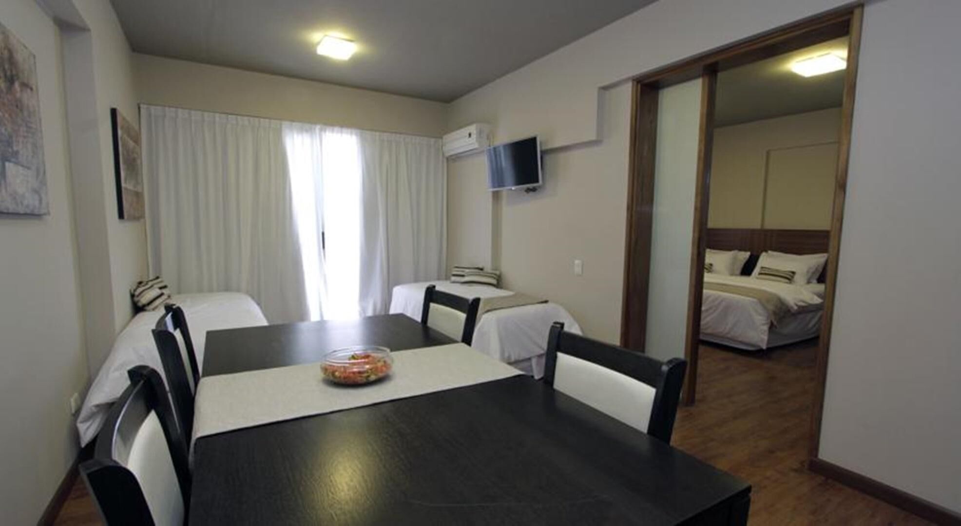 Une chambre d’hôtel équipée d’une table à manger, de chaises, d’un téléviseur fixé au mur et d’un lit avec une tête de lit en bois.