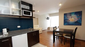 A modern kitchen with dark cabinetry, a white dishwasher, and a granite countertop. There is a dining area with a wooden table and chairs, and a painting on the wall.