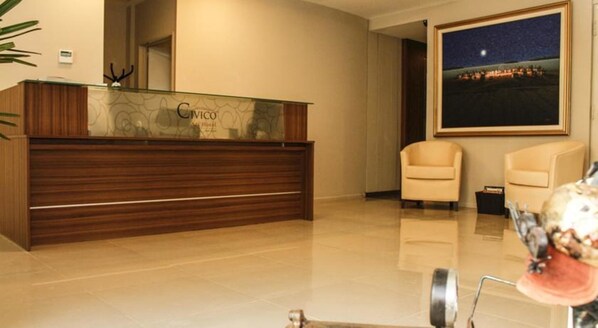 Un mostrador de recepción con encimera de vidrio y base de madera, dos sillones color beige y un cuadro con una escena nocturna en la pared.