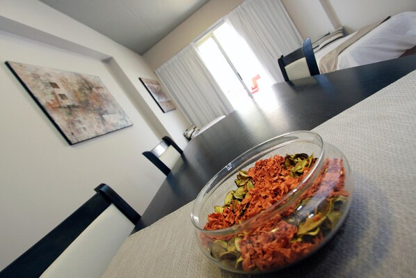 A dining table with a glass bowl containing dried herbs, a painting on the wall, and a bed in the background.