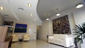 A modern hotel lobby with a reception desk, two armchairs, a sofa, a large abstract painting, and recessed ceiling lighting.
