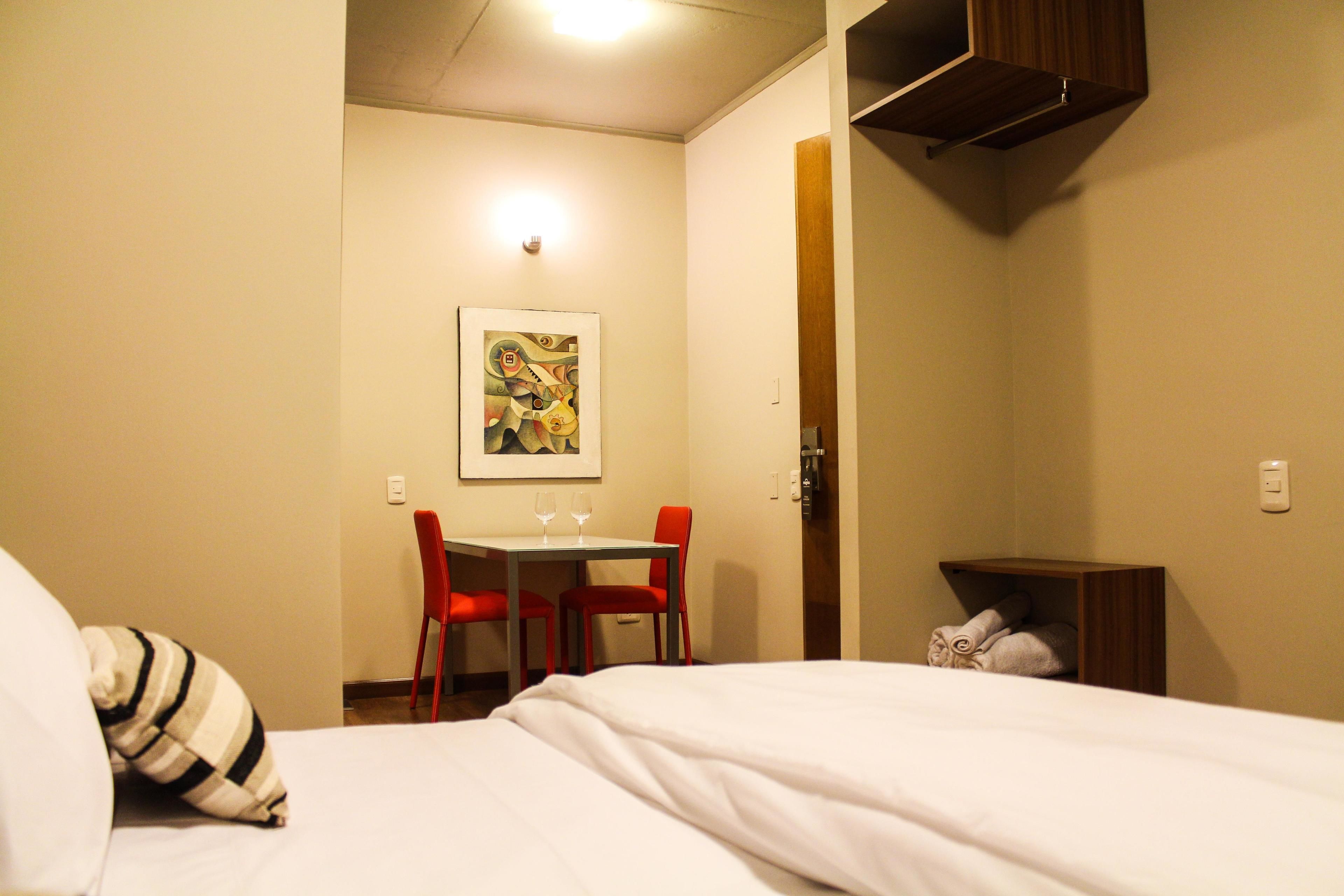 A hotel room with a bed, a desk with two chairs, a painting on the wall, and a small wooden cabinet.