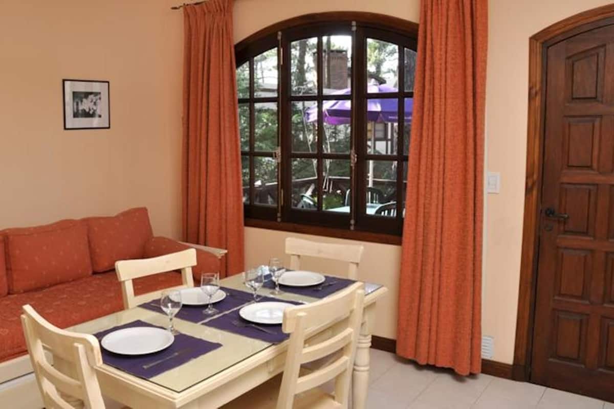 Um quarto aconchegante com um sofá, uma mesa de jantar posta para dois e uma janela com cortinas.