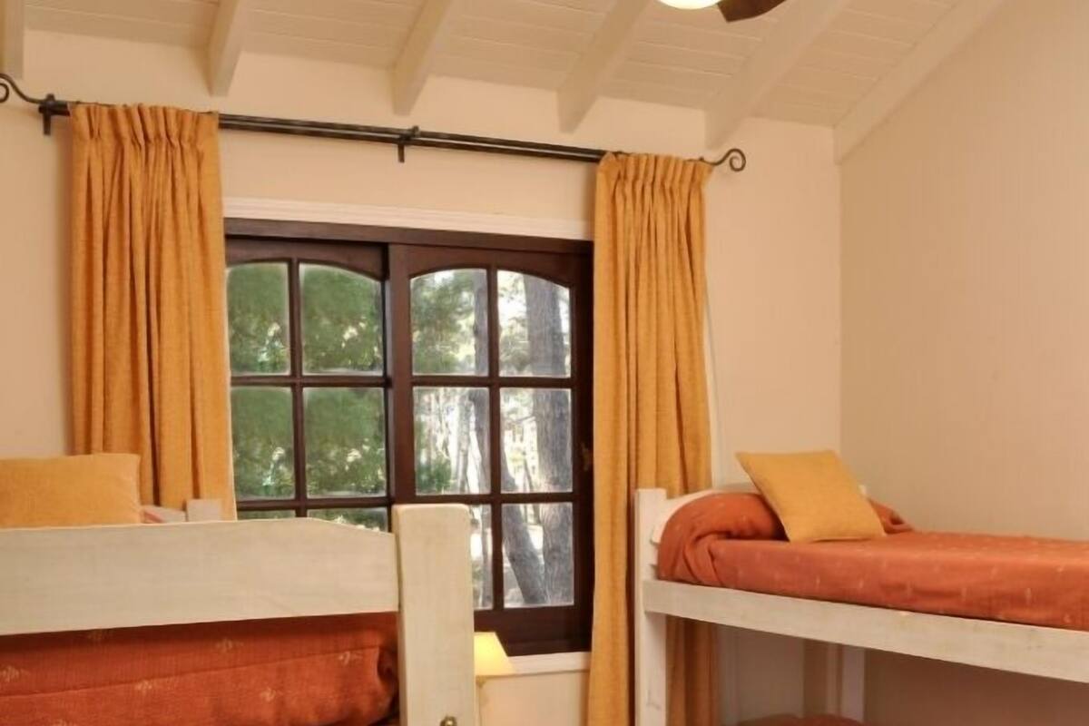 Quarto de dormitório com beliches, janela com cortinas, ventilador de teto e cômoda branca pequena.