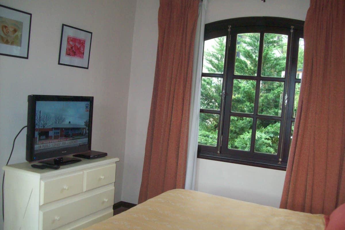 Um quarto com uma cama, cômoda, televisão sobre a cômoda e uma janela com cortinas.