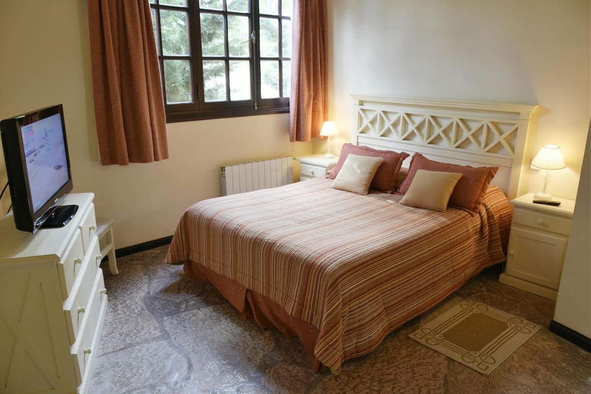 Quarto com cama, duas mesas de cabeceira, televisão sobre cômoda e janela com cortinas.