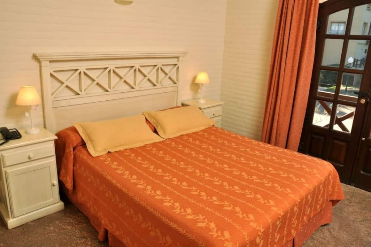 Uma cama com colcha laranja estampada com flores, cabeceira branca com design geométrico, duas luminárias de cabeceira e uma porta de madeira com painel de vidro.