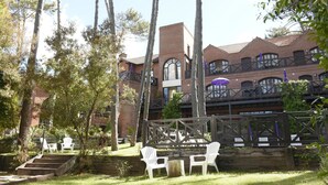 Un bâtiment à la façade en briques, doté de grandes fenêtres et d’un balcon agrémenté de parasols. On y trouve des espaces de détente en plein air avec des chaises blanches et une terrasse en bois.