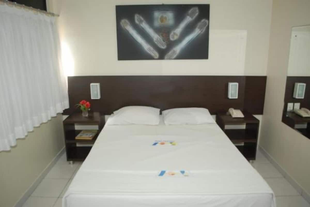 Habitación de hotel con una cama grande, dos mesitas de noche con lámparas, una obra de arte en la pared y una ventana con cortinas.