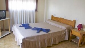 Una cama individual con cabecera de madera, una mesita de noche con lámpara, y un televisor sobre un soporte.