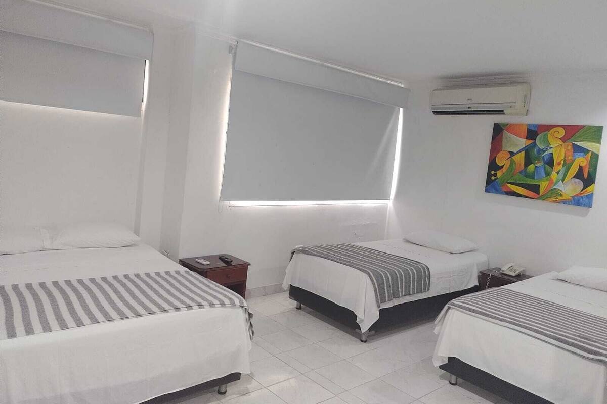 A hotel room with two single beds, a window with curtains, an air conditioning unit, and a colorful abstract painting on the wall.