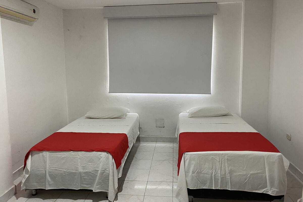 A small, clean room with two single beds, white walls, a window, and a white air conditioning unit.