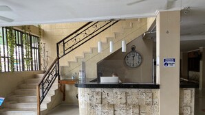 Un hall d’hôtel avec un comptoir de réception en marbre, un escalier doté d’une rampe métallique, une horloge au mur et un panneau indiquant qu’il est possible de boire l’eau du robinet.