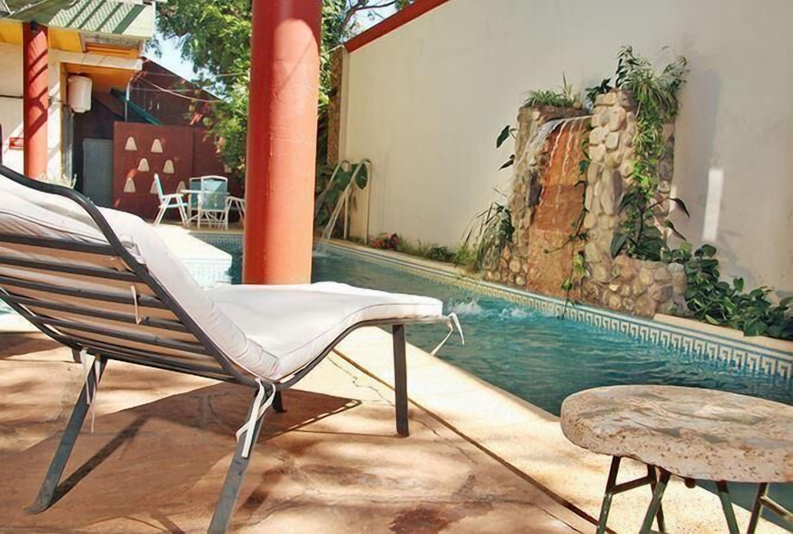 Zona de piscina con una silla de descanso, una mesita y un muro de piedra con una cascada.