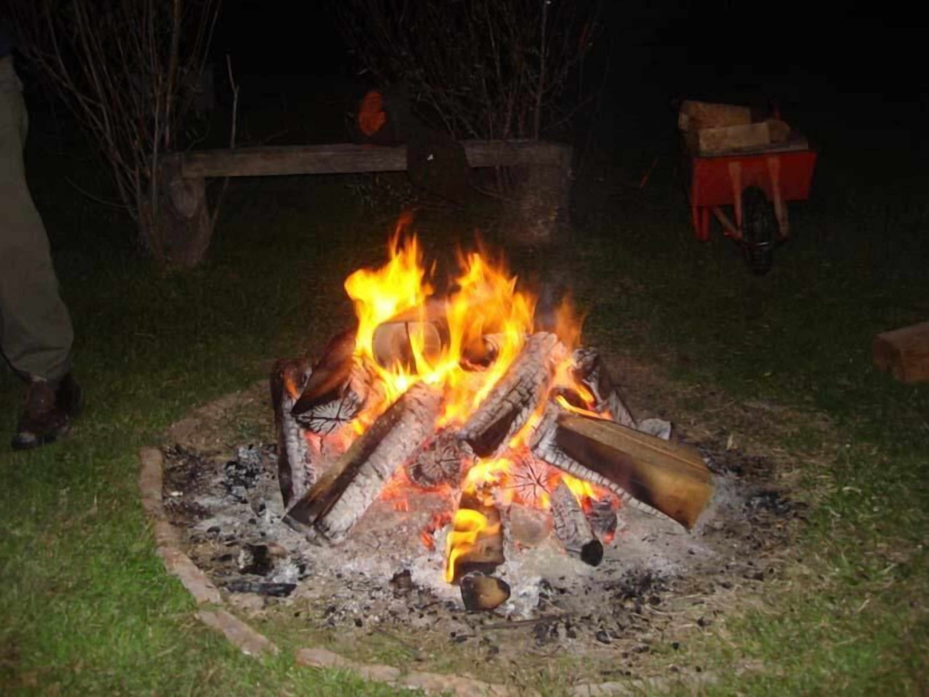 Una fogata con leña y madera ardiendo, rodeada de hierba y una valla de madera.