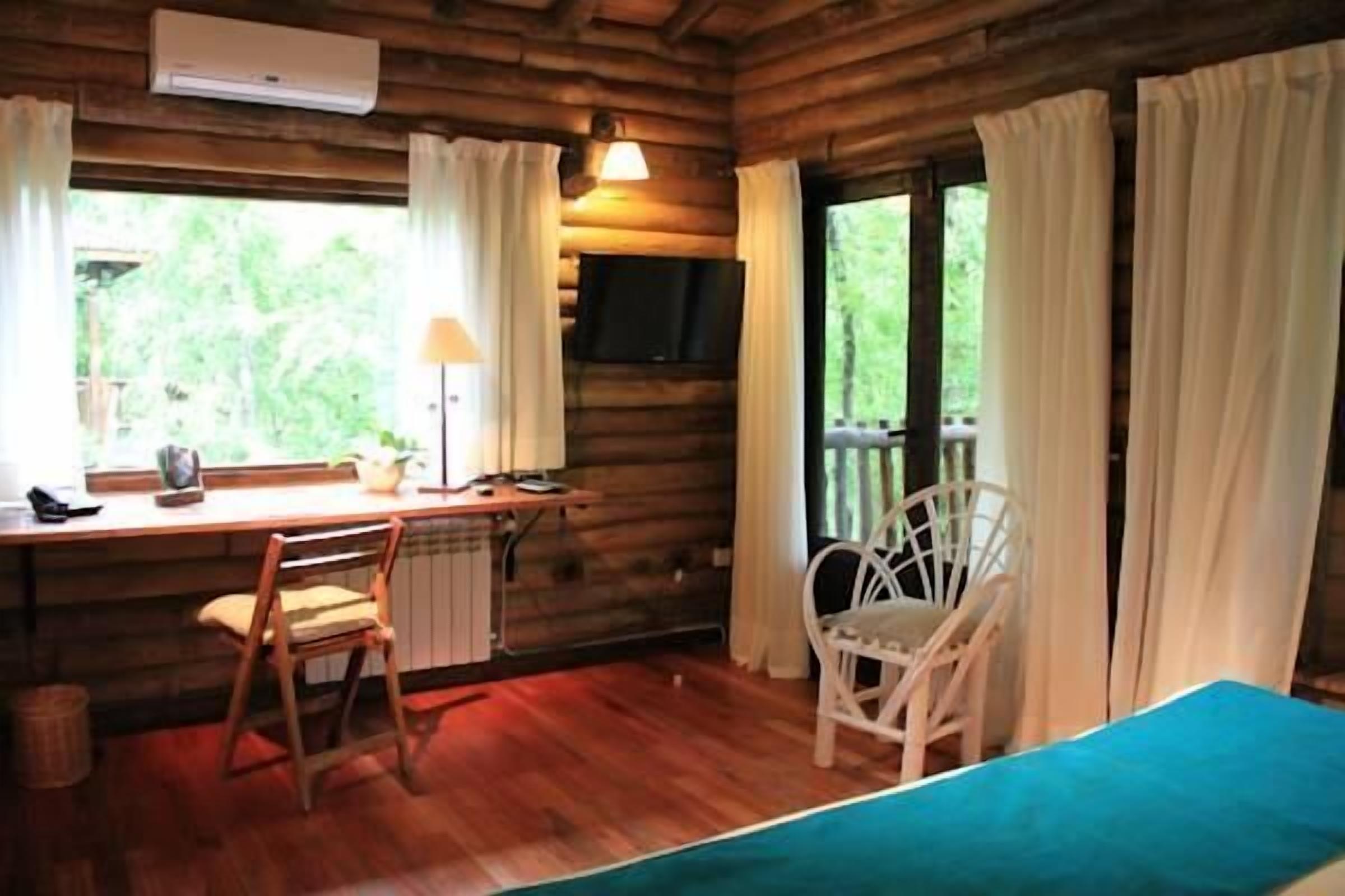 Ein Zimmer einer Holzhütte mit einem Bett, einem Schreibtisch mit Stuhl, einem Fernseher und einem Fenster mit Vorhängen.