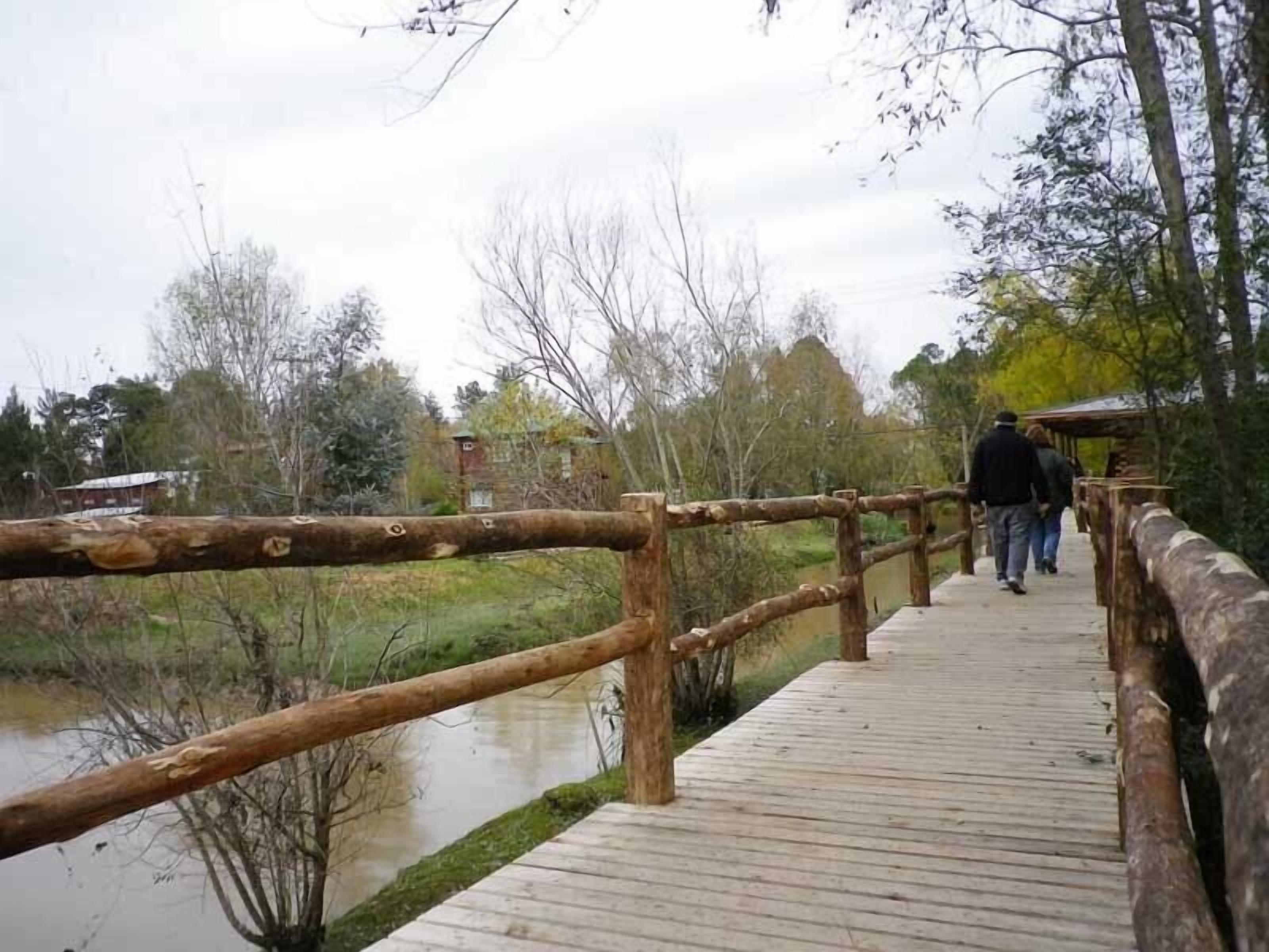 A wooden boardwalk over a body of water, surrounded by trees and a building in the background.