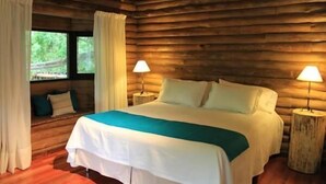 A log cabin room with a bed, two bedside lamps, a window with curtains, and a view of greenery outside. - Amarran Sancho (Tigre)