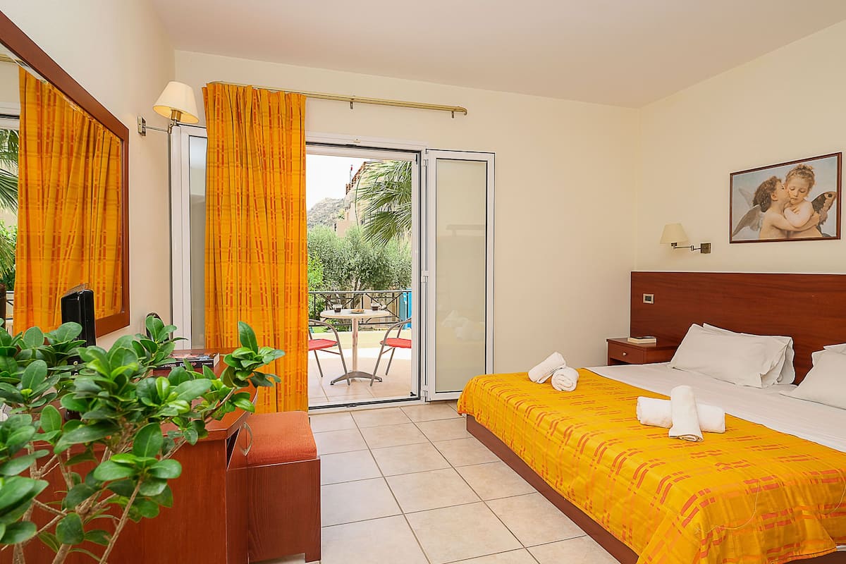 A hotel room with a bed, a desk, a chair, a small table, a plant, and a view of the outside balcony.
