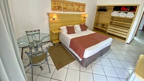Una habitación de hotel con una cama, dos mesitas de noche con lámparas, una mesa de vidrio con una silla, un armario de madera y piso de baldosas.