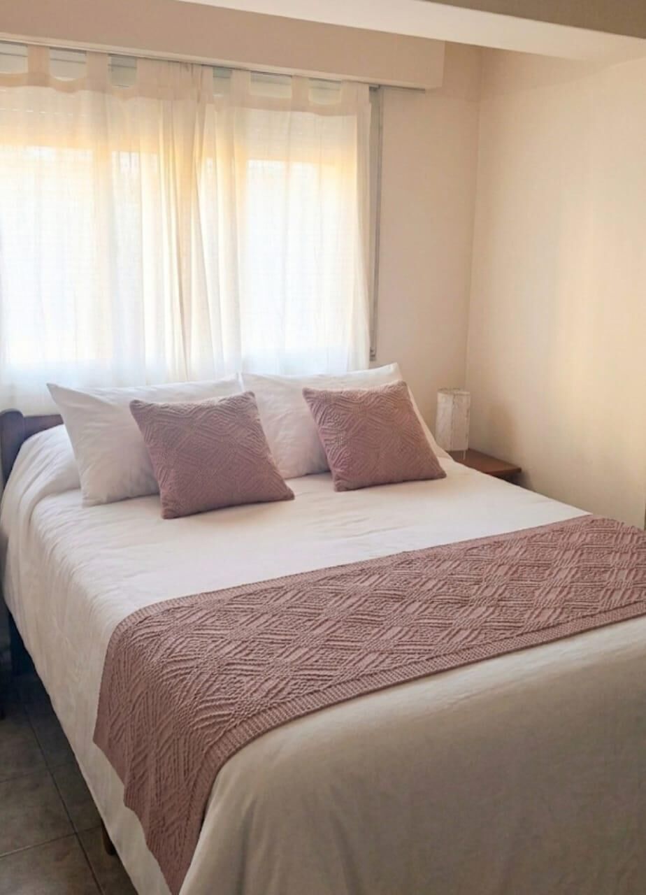 A neatly made bed with a patterned blanket, a bedside table with a lamp, and sheer curtains.