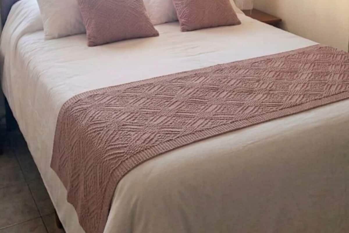 Una cama bien hecha con un edredón estampado, una mesita de noche con una lámpara y cortinas de gasa.