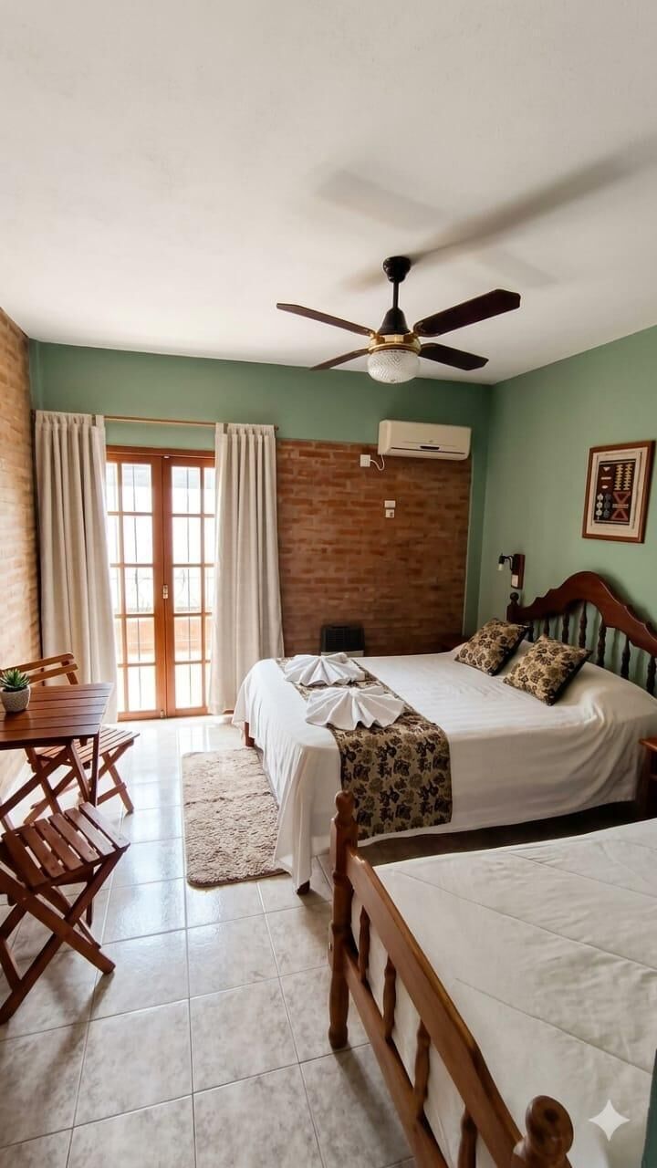 A bedroom with a bed, a ceiling fan, a wooden headboard, a table with chairs, and a window with curtains.