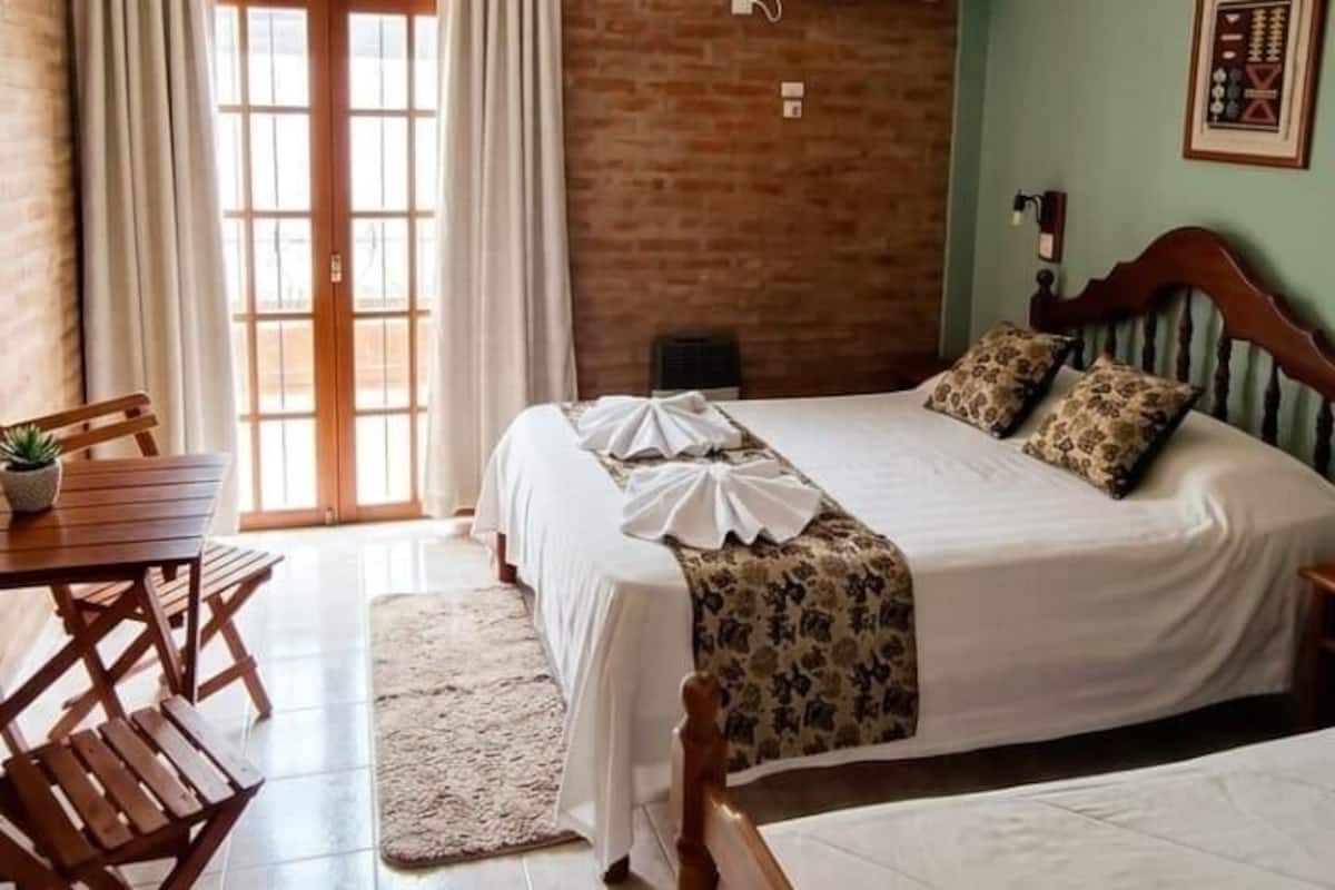 Un dormitorio con una cama, un ventilador de techo, un cabecero de madera, una mesa con sillas y una ventana con cortinas.