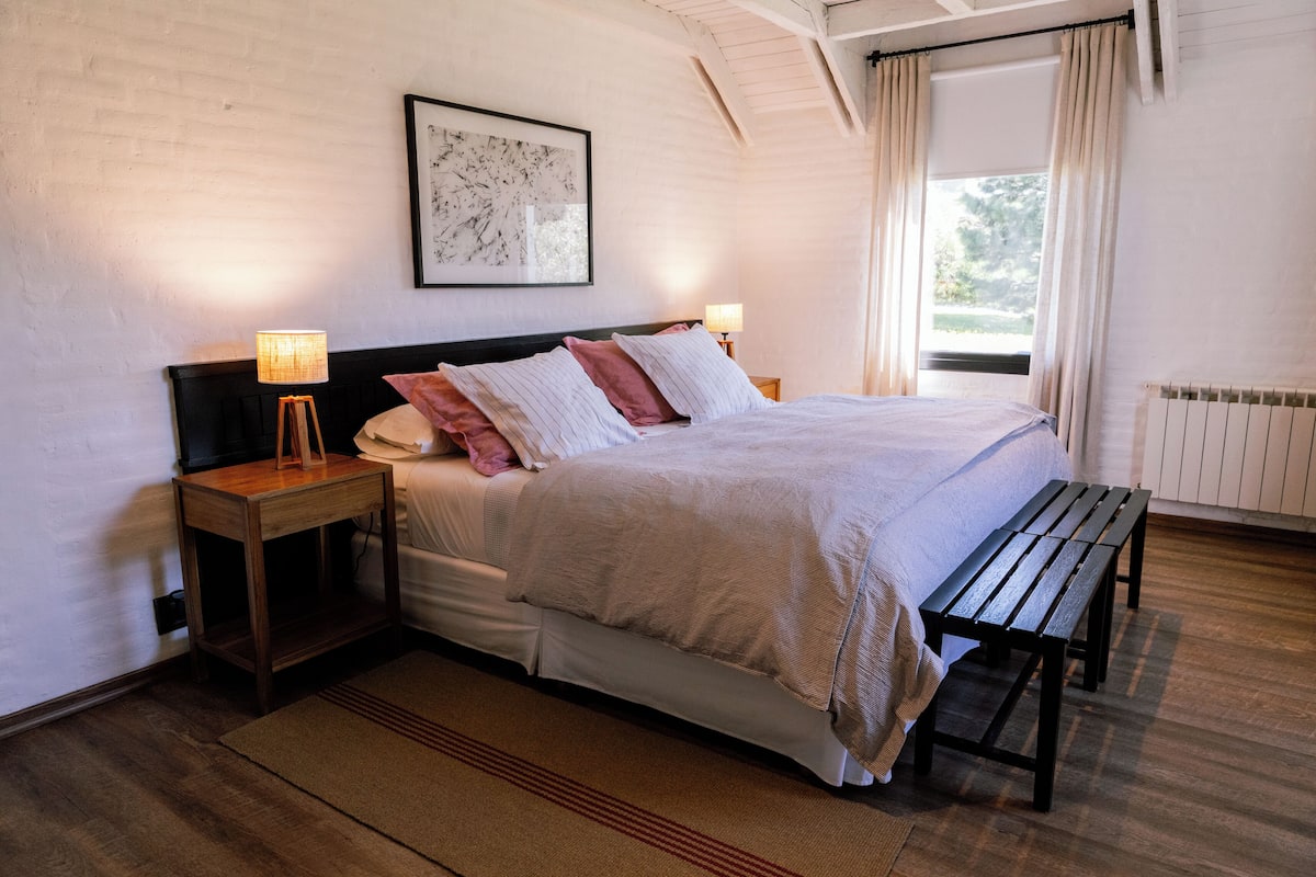 Un dormitorio ordenado con cama, mesitas de noche, un banco y una ventana con cortinas.