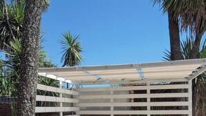 A person sitting on a white cushioned bench with a white metal frame, reading a book, surrounded by greenery and palm trees. - Cabañas la Aldea (Villa Carlos Paz)