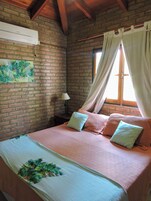 Un dormitorio con pared de ladrillo, una cama con colcha estampada, dos almohadas, una mesita de noche con lámpara y una ventana con cortinas.