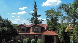 A two-story house with a tiled roof, a front yard with a paved walkway, and a large tree in the background. - Cabañas la Aldea (Villa Carlos Paz)