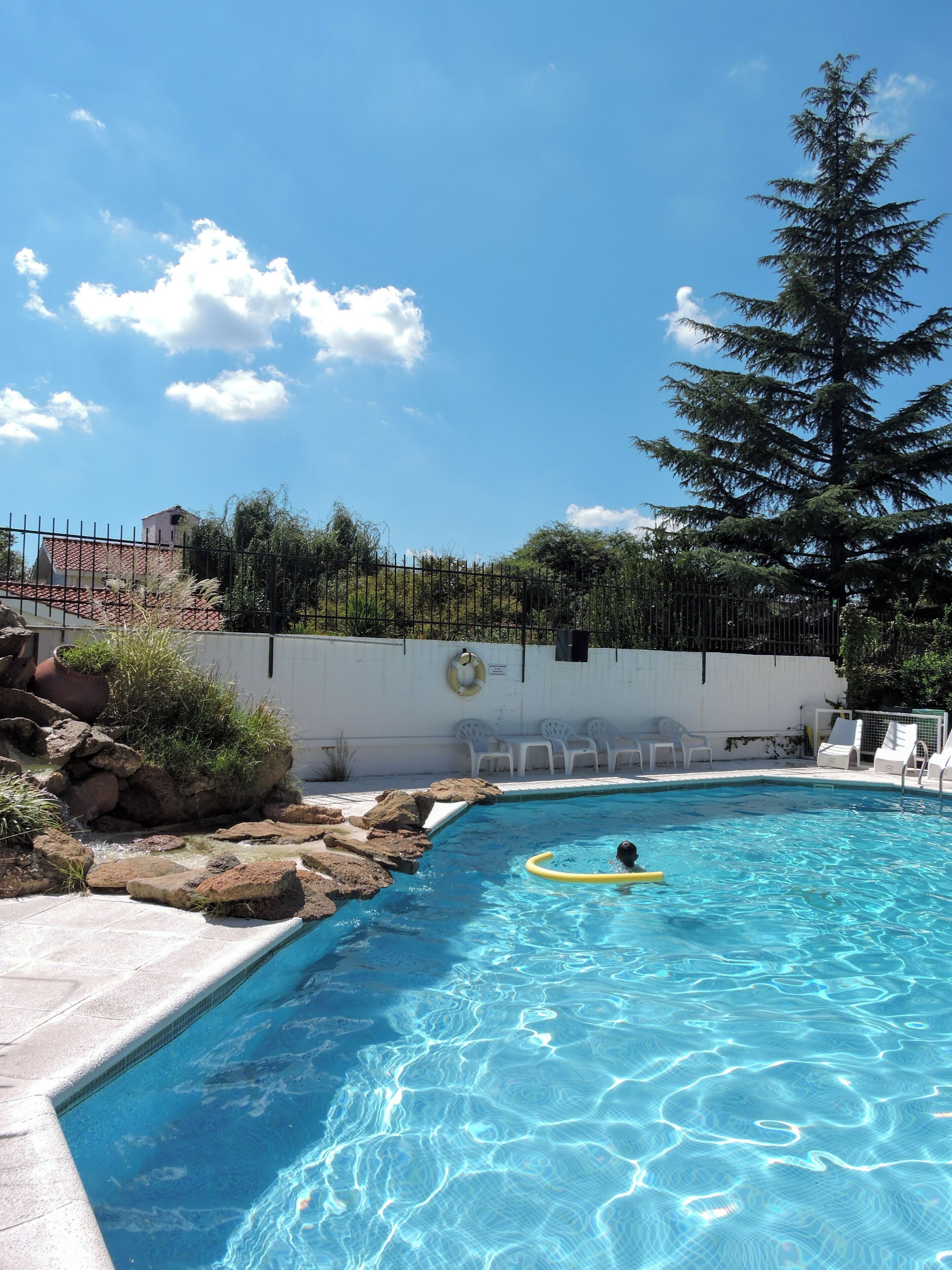 Una piscina con agua azul cristalina, rodeada de rocas y plantas, y una cerca blanca con un flotador salvavidas.
