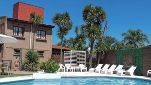 A swimming pool with lounge chairs, palm trees, and a brick building. - Cabañas la Aldea (Villa Carlos Paz)