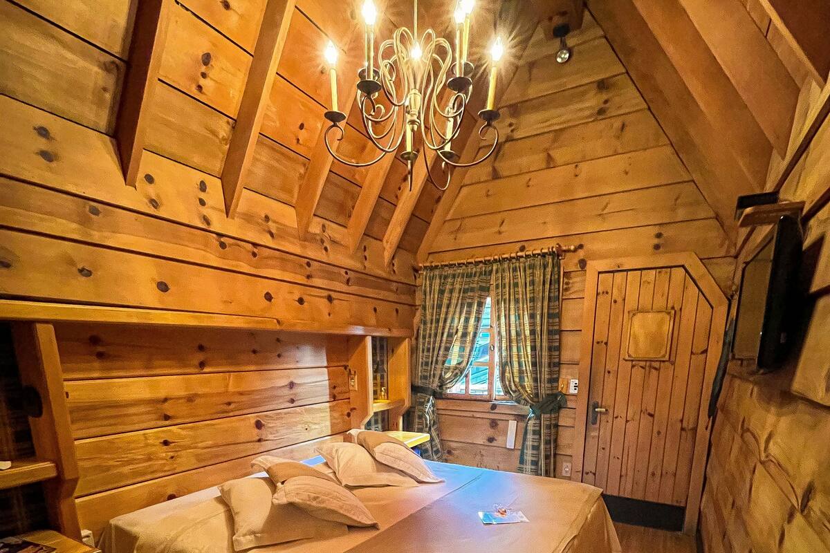 A wooden cabin bedroom with a bed, a nightstand, a chandelier, and a window with curtains.