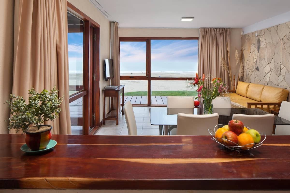 A room with a wooden table, a bowl of fruit, a potted plant, a sofa, and a view of the beach.