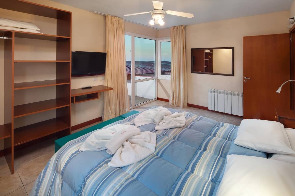 A hotel room with a bed, a TV, a wardrobe, a balcony with curtains, and a ceiling fan.
