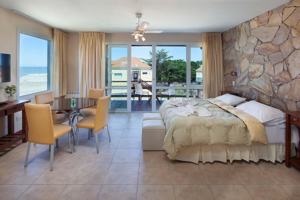 A hotel room with a bed, a dining table with chairs, a TV, and a balcony with a view of the beach.