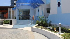 A modern building with a blue awning over the entrance, a staircase leading to the entrance, and a small garden area with plants and a white bollard.